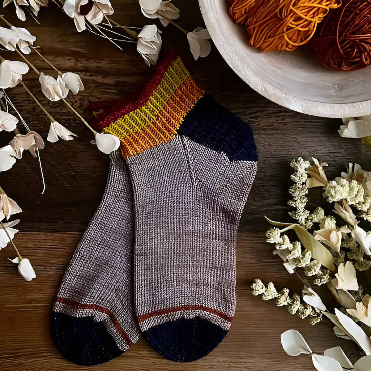 Pair of gray socks with black toes and red accents on a wooden surface with yarn and flowers.