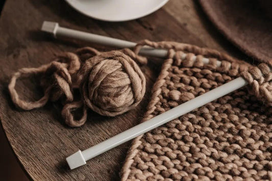 brown yarn, knitting needles, and a chunky knitted project on a wooden table