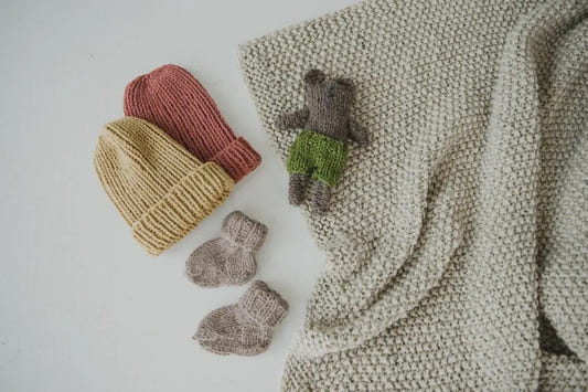 hand-knit baby hats, a toy bear, and booties beside a cream knitted baby blanket