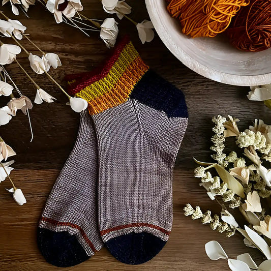 Pair of gray socks with black toes and red accents on a wooden surface with yarn and flowers.