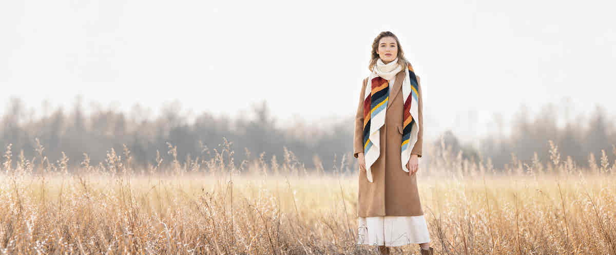 Woman in a camel coat wearing the Gresham Wrap, a long striped shawl knit in Blue Sky Fibers Woolstok Worsted, standing in a field of dry autumn grass