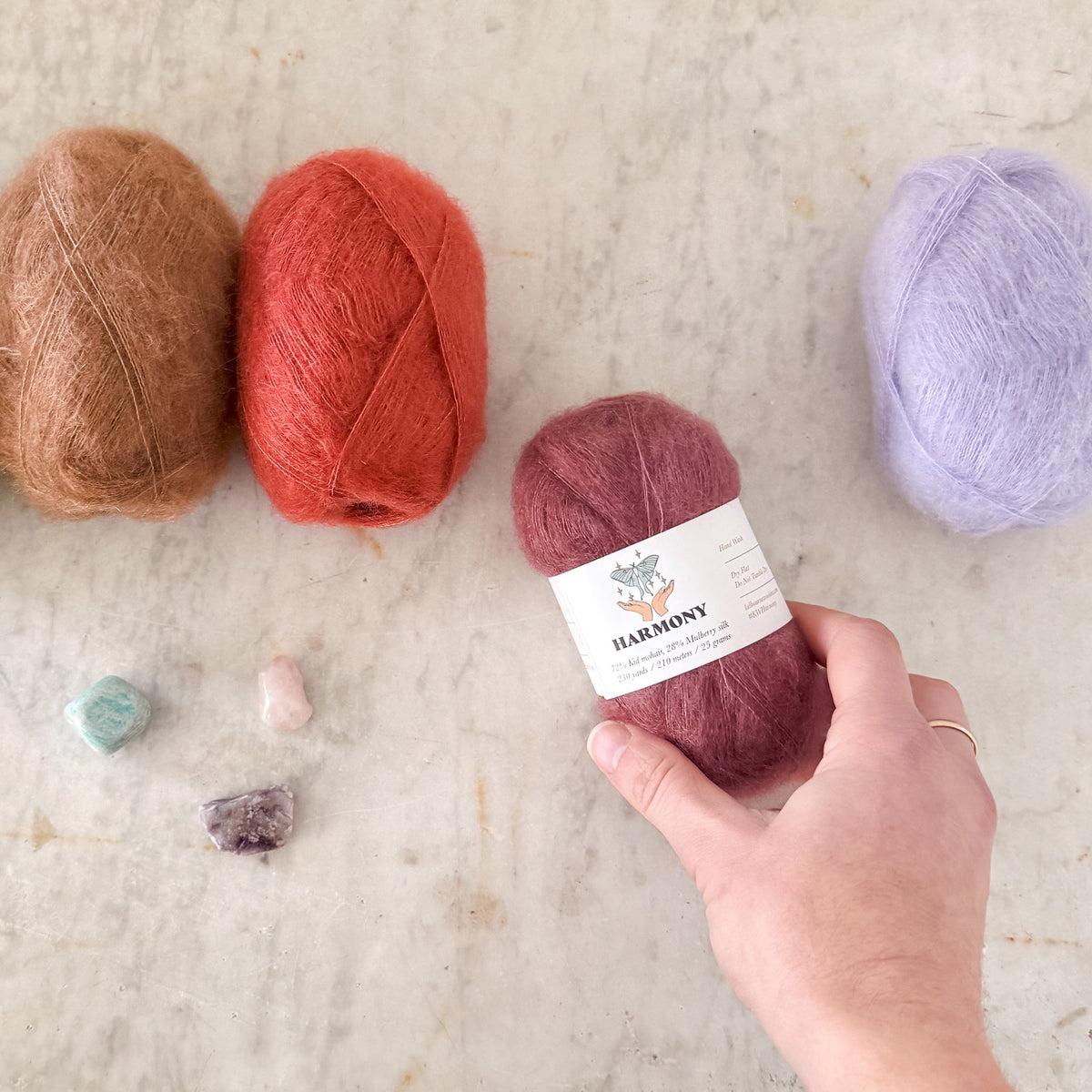 A ball of Kelbourne Woolens Harmony Lace Yarn held in hand beside three more colours of the kid mohair and silk lace yarn
