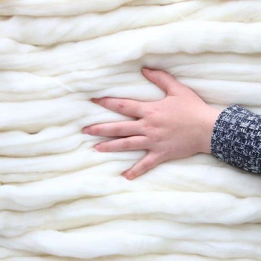 A hand resting on thick coils of undyed natural white Merino wool top, showing the scale and softness of the fiber