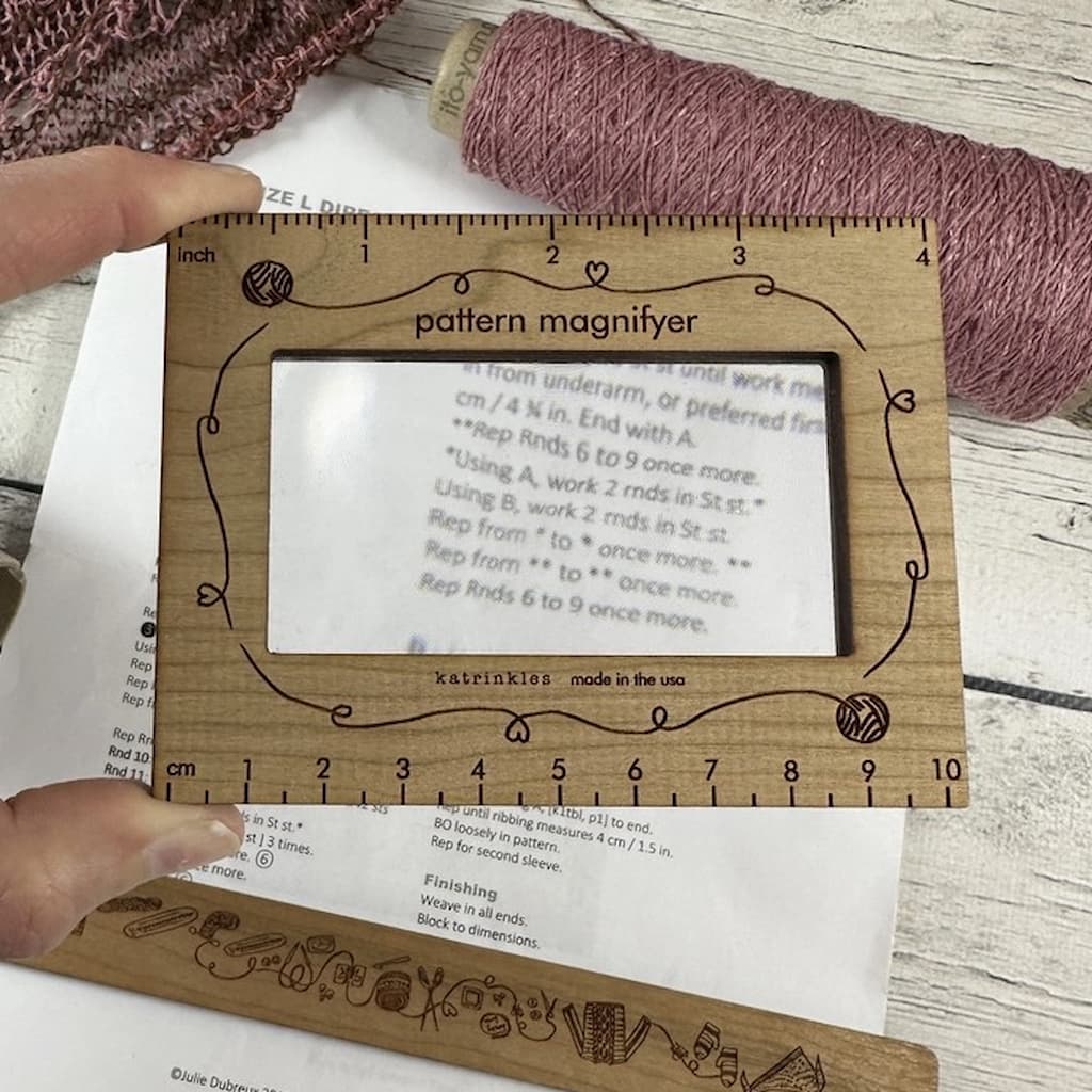 A pattern magnifier tool with a wooden frame and clear plastic lens, placed over a knitting pattern next to a ball of yarn.