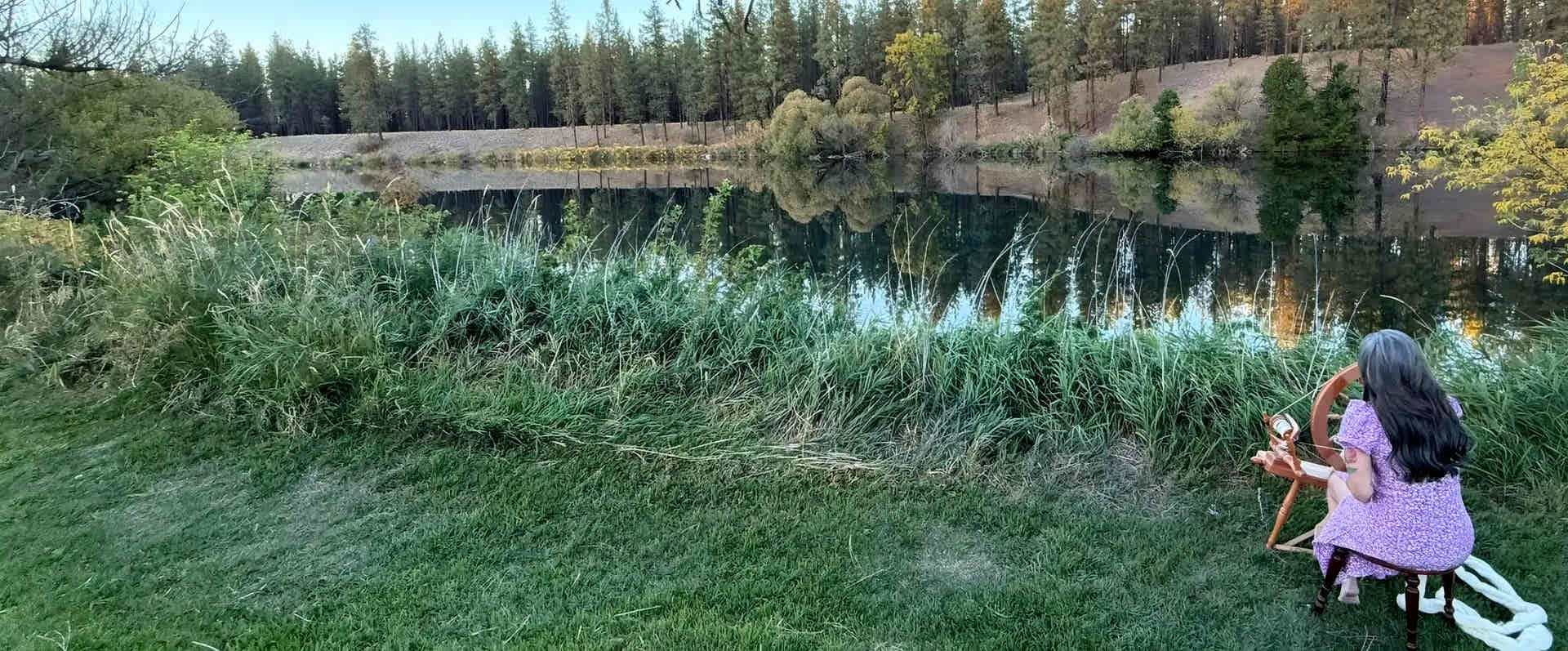 Sara sitting by the Spokane River surrounded by trees and spinning with a wheel