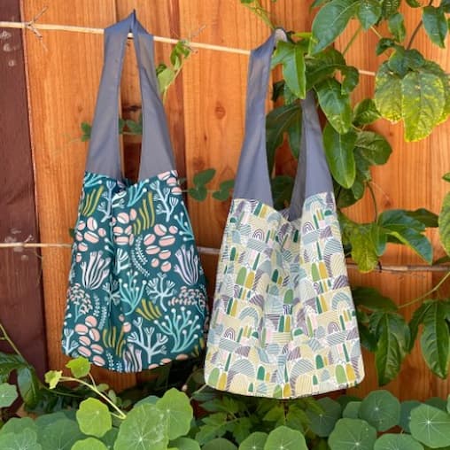 2 Flip & Tumble tote bags hanging with green foliage in front of a wooden fence.