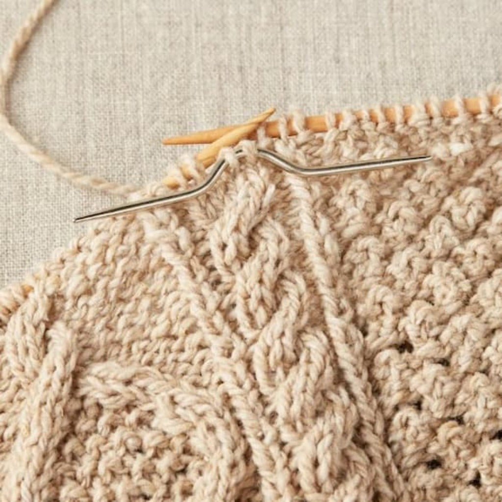 Silver cable needle holding cable stitches on a cream colored cabled knitting piece