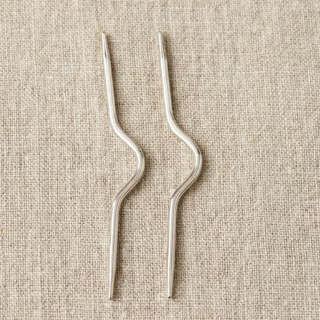 Two silver cable needles laying side by side on a linen cloth
