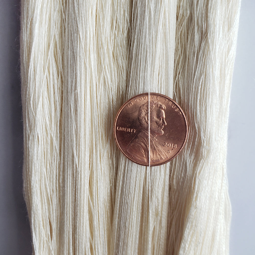 A close up of Paradise Fibers 140/2 Undyed Spun Silk Cobweb Yarn with a penny for size.