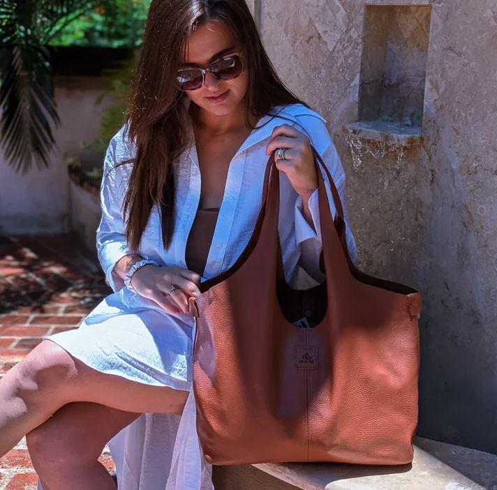 Brunette Woman sitting on a bench holding a tan colored Boundless leather bag in the color Almond.