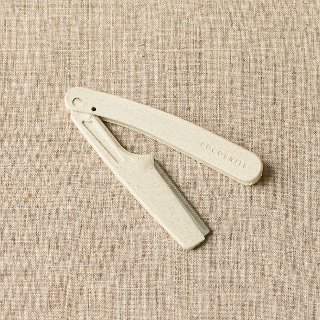 A white CocoKnits Fuzz Off Comb laid on a textured fabric surface.