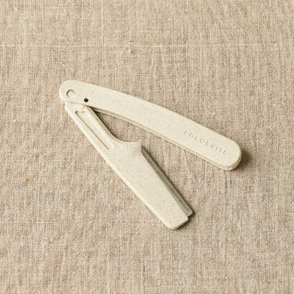 A white CocoKnits Fuzz Off Comb laid on a textured fabric surface.
