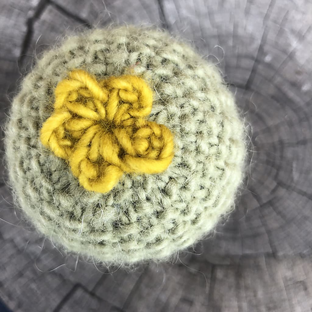 Top view of a small, round, light green knitted cactus with a yellow crocheted flower on top. 