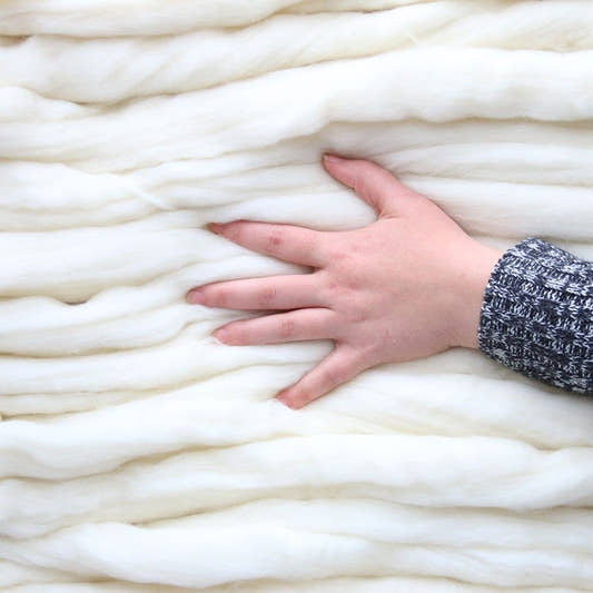 A hand resting on thick coils of undyed natural white Merino wool top, showing the scale and softness of the fiber